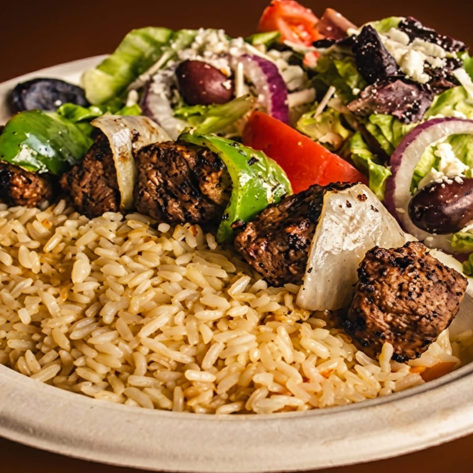 Plate of grilled meat skewers with peppers and onions, seasoned rice, and Greek salad with lettuce, olives, tomatoes, and feta.