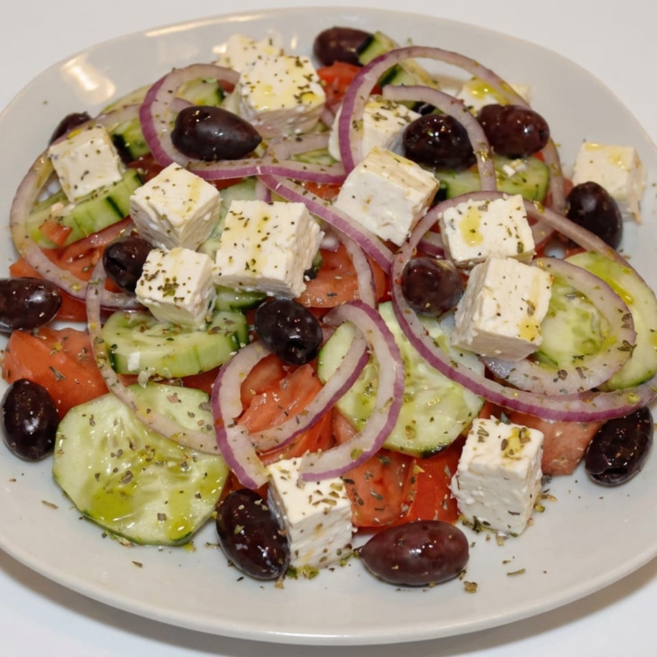 Greek salad with tomatoes, cucumbers, red onions, feta cheese, Kalamata olives, and herbs on a white plate.